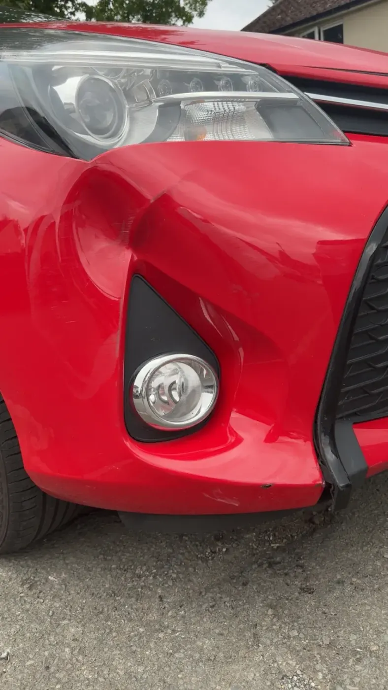 Before: Red Toyota Yaris with a large dent in the front bumper next to the fog light
